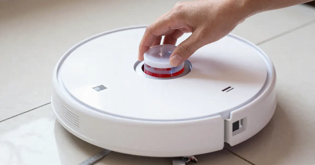 Illustration of a hand resting on top of a generic white robot vacuum mop on a light tiled floor