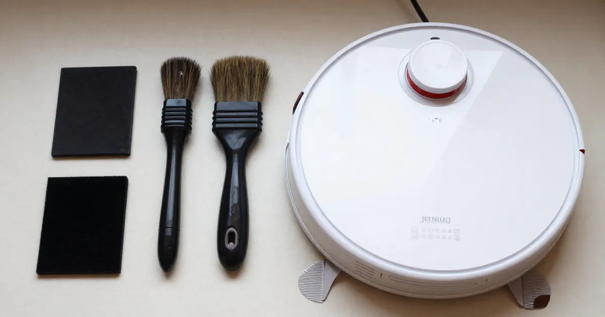 Illustration of a generic white robot vacuum next to two small brushes and two dark filter pads on a white surface